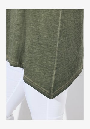 Haut vert olive avec une texture douce et un ourlet arrondi, présentant des détails de couture légers. Porté avec un pantalon blanc moulant.