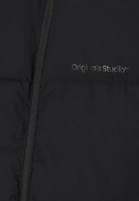 Chaqueta negra acolchada con cierre de cremallera y una textura suave. Presenta el logotipo "Originals Studio" en un sutil color gris.