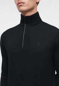 Homme portant un pull en maille noire avec un col montant et une courte fermeture éclair à l'avant, laissant visible le haut de la poitrine et la zone du cou.