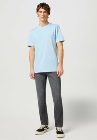 Camiseta de algodón azul claro con cuello redondo y mangas cortas, combinada con jeans grises y zapatos negros con suelas blancas.