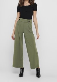 Olivově zelené kalhoty s širokými nohavicemi a vysokým pasem, se dvěma předními kapsami a černými knoflíky. V kombinaci s přiléhavým černým žebrovaným topem a černými botami.