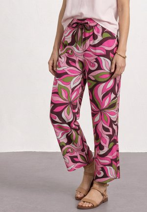 Femme portant un pantalon à jambes larges avec un motif floral rose, marron et vert, des sandales beiges et un top sans manches rose clair.