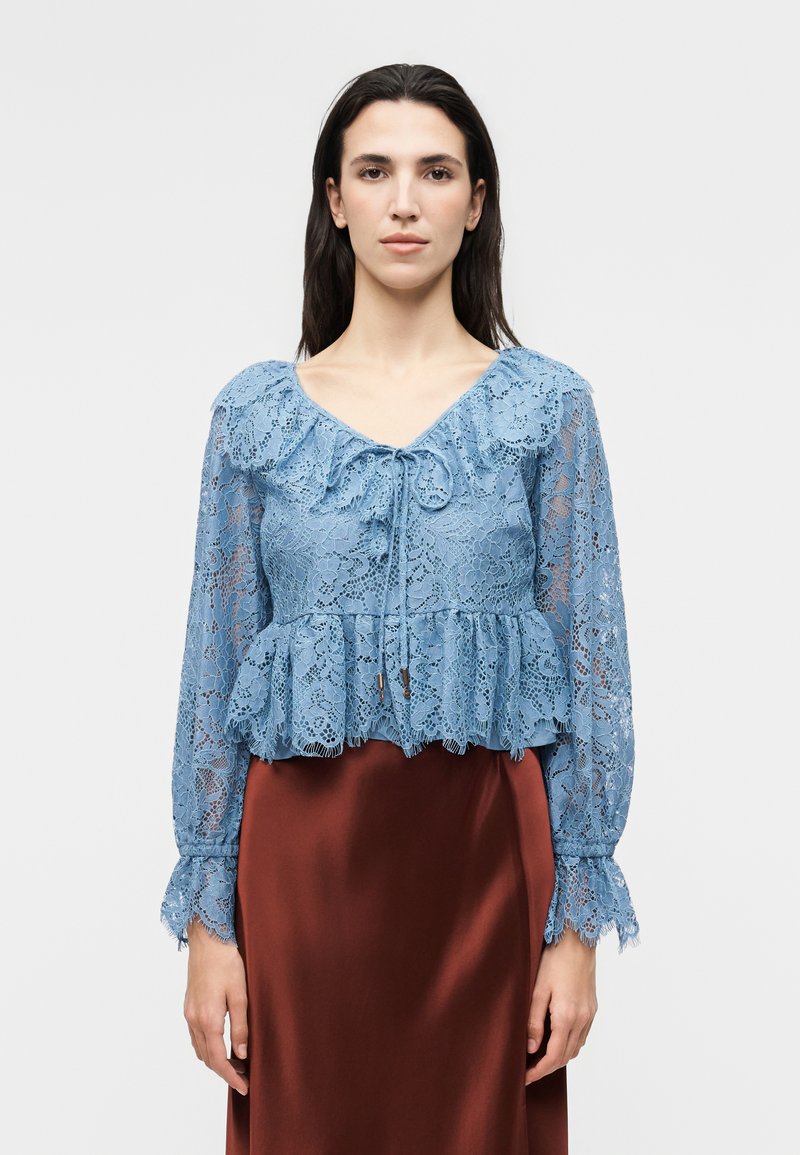 Γυναίκα που φορά μπλε δαντελένιο μπλουζάκι με μακριά μανίκια, με βολάν στο τελείωμα και στον λαιμό, δεμένο με φιόγκο, σε συνδυασμό με καφέ σατινέ φούστα.