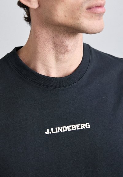 Juoda medvilnės marškinėliai su apvaliu kaklu; priešinėje pusėje raudona spauda "J.LINDEBERG". Lygus paviršius, prigludęs dizainas.