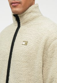 Muškarac nosi bež teksturiranu zip-up flis jaknu s crnim patentnim zatvaračem i malim pravokutnim logotipom na prsima.