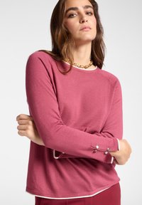 Maglietta lunga rosa con collo rotondo e rifiniture bianche. Presenta dettagli con bottoni sui polsini e un tessuto morbido e testurizzato.