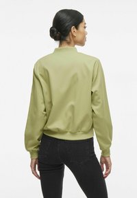 Giacca bomber verde chiaro con colletto a coste, maniche lunghe e polsini elasticizzati; texture liscia e vita aderente; indossata con jeans neri.