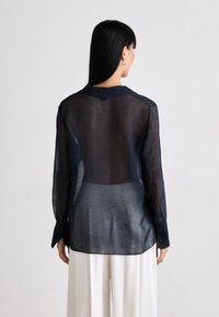 Mulher vestindo uma blusa azul marinho transparente de mangas longas e calças brancas de pernas largas, de pé de costas contra um fundo simples.