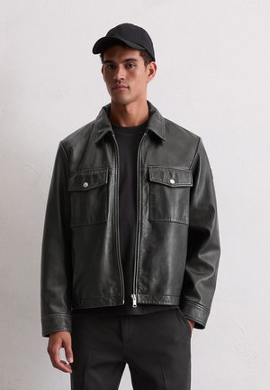 Black faux leather jacket with a collar, two chest pockets, and silver snap buttons; worn over a black top, featuring a smooth texture.