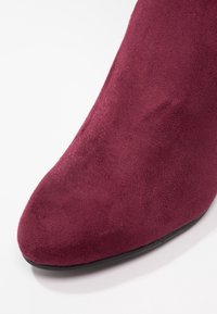Gros plan sur une chaussure pointue en suède bordeaux avec une semelle basse noire sur fond blanc.