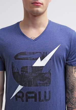 Niebieska koszulka t-shirt z dekoltem w serek wykonana z miękkiego materiału, z graficznym nadrukiem noża i stylizowanym tekstem. Podkreślona białym wzorem błyskawicy.