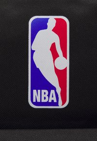 Logo de la NBA représentant un joueur en silhouette dribblant un ballon de basket, en blanc, bleu et rouge sur un fond noir texturé.