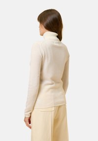 Krämfärgad turtlenecktröja i mjukt material, åtsittande silhuett, med ribbad krage och långa ärmar, stylad med ljusa byxor.