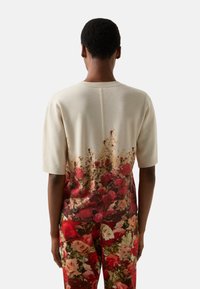 Beżowy sweter dziany z kwiatowym wzorem w odcieniach czerwieni i zieleni przy dolnym brzegu. Krótkie rękawy i okrągły dekolt. Miękka tekstura, luźny fason.