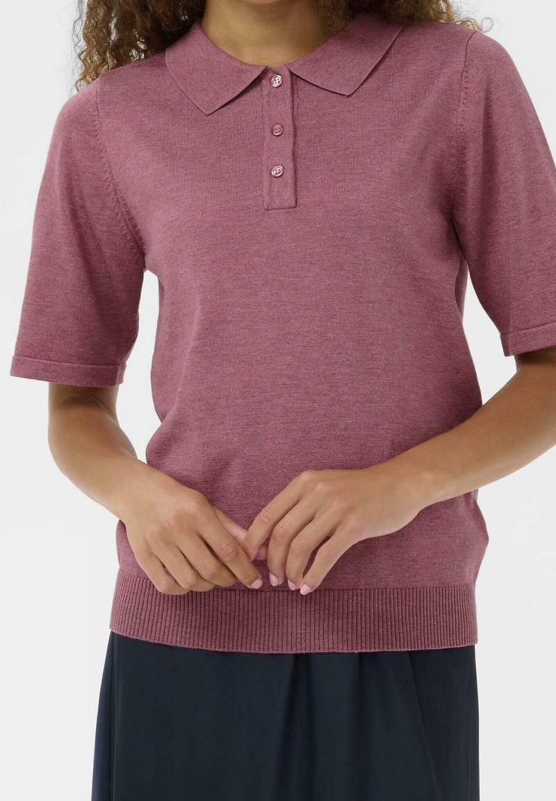Femme portant un polo en maille rose poudré à manches courtes avec trois boutons et ourlet côtelé, mains jointes devant, jupe noire en dessous.