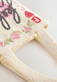 Crèmekleurige gekralen handtas met roze bloemenborduursel, groene bladeren, zwarte letters en rode hartvormige kralendetails.