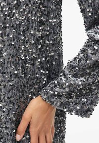 Sudraba krāsas audums ar teksturētu virsmu, rotāts ar spīguļiem. Piedurknes gals beidzas ar aproci. Spīguļi atstaro gaismu, radot mirdzošu efektu.