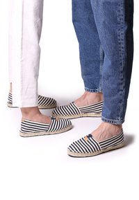 Prúžkované espadrilky s námorníkovou a bielou tkaninou, so šúpkovými podrážkami a zaoblenou špičkou. Dve dvojice obuté s ležérnymi džínsami a bielymi nohavicami.