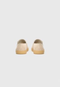 Un par de zapatos de ante beige sin cordones con suelas de goma, vistos desde atrás sobre un fondo gris claro.