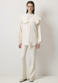 Blusa crema con cuello grande y volantes, combinada con pantalones crema ajustados. La tela parece suave con una textura lisa.