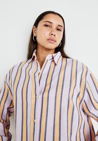 Chemise à boutons en tissu doux, avec des rayures verticales lavande, orange et bleu marine. Col classique et boutons clairs.