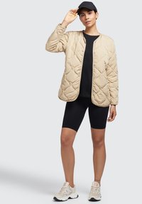 Beige gewatteerde jas met drukknoopsluitingen, gedragen over een zwart top en ribbelachtige zwarte shorts, gecombineerd met beige en witte sneakers.
