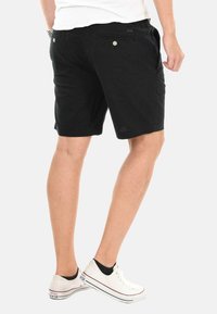 Schwarze Baumwollshorts mit Seitentaschen und zwei hinteren Knöpfen. Getragen mit weißen Sneakers, die eine entspannte Passform und ein lässiges Design zeigen.