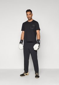 Svart t-skjorte med korte ermer og teksturert lomme, svarte bukser, hvite boksehansker med svarte detaljer, og svarte joggesko med beige såler.
