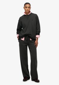 Sort oversized sweatshirt med rund halsudskæring, kombineret med en lyserød skjorte under og løstsiddende sorte bukser. Brune sko.