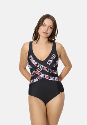 Mujer con traje de baño negro de una pieza con paneles transparentes de flores multicolores, de pie con las manos detrás de la espalda sobre un fondo liso.