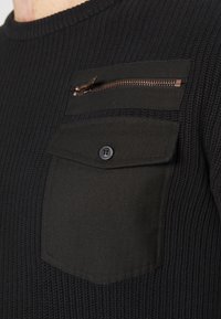 Schwarzer Strickpullover mit gerippter Textur, ausgestattet mit einer Brusttasche mit Knopf und einer Reißverschlusstasche, beide aus einem kontrastierenden Stoff.