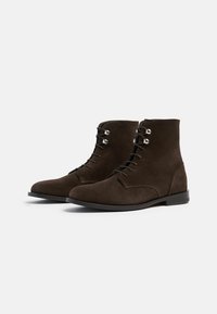 Botines de ante marrón hasta el tobillo con punta redondeada, sistema de cordones y ojetes plateados. Diseño minimalista con una suela de cuero lisa.