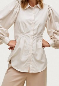 Ljust beige puffärmad skjorta med knappar och figurnära midja, slät textur och veckdetaljer fram och bak.