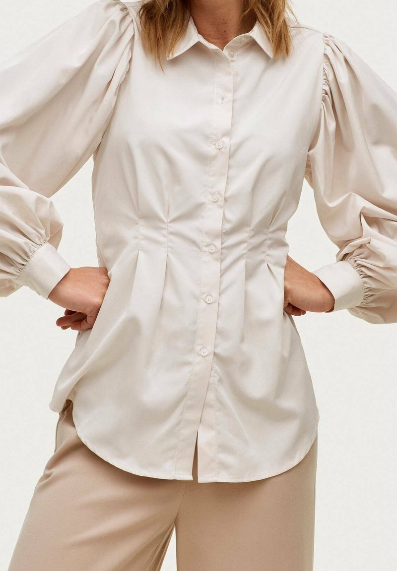 Ljust beige puffärmad skjorta med knappar och figurnära midja, slät textur och veckdetaljer fram och bak.