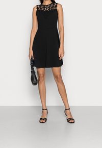 Svart ärmlös klänning med spetsöverlägg vid halsringningen, vid kjoldel och slät textur. Matchas med svarta sandaler med remmar och en clutch.