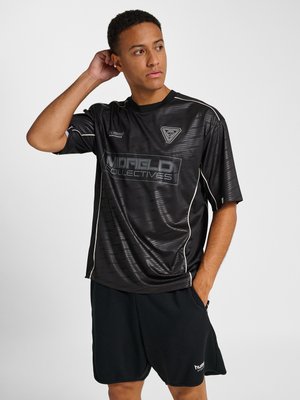 Junger Mann in schwarzer Sportbekleidung mit dem Logo „Midfield Collectives“ auf dem Shirt, der mit einer Hand hinter dem Kopf und der anderen in der Tasche steht.