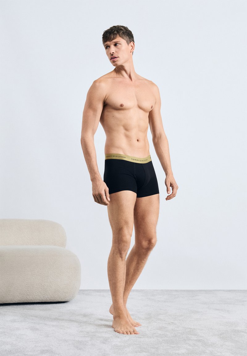 Sorte bomuldsboxershorts med en gul logo-linning. Modellen står på et lys gråt tæppe og viser en minimalistisk indretning.
