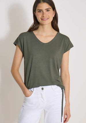 Jeune femme portant une chemise à manches courtes vert foncé avec un lien sur le côté et un pantalon blanc, souriante avec une main dans la poche sur fond uni.