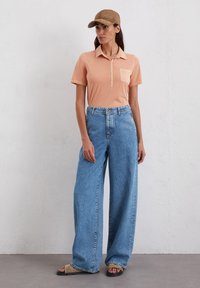 Perzikkleurig poloshirt met een kraag en knoopdetail, gecombineerd met wijd-uitlopende lichtblauwe denimjeans. Een tankleurige pet maakt de outfit compleet.