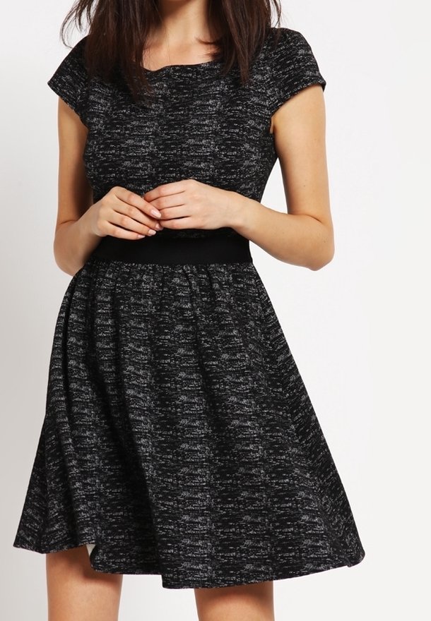 Femme portant une robe noire et grise texturée à manches courtes, longueur genou, avec une ceinture cintrée, mains jointes à la taille.