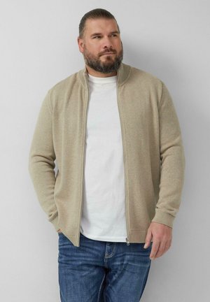 Brodaty mężczyzna z zaczesanymi do tyłu włosami, ubrany w beżowy sweter na zamek błyskawiczny na białej koszulce i niebieskich dżinsach, stojący na tle gładkiego tła.