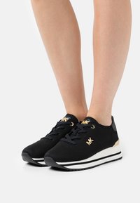 Sorte tekstilsko joggesko med gullfunn og hvit og svart stripete såle, teksturert overdel og rund tå. Snørt med synlig merkevare.
