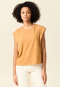 Top senza maniche giallo senape con una vestibilità comoda, in morbido cotone, colletto rotondo e design delle spalle senza cuciture. Abbinato a pantaloni bianco sporco.