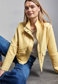 Veste en tissu jaune avec fermeture éclair à l'avant, col snap et deux poches avant, associée à un jean en denim bleu. Texture lisse, design décontracté.
