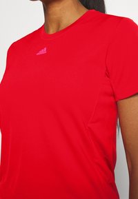 Camiseta deportiva roja de manga corta hecha de una tela suave. Presenta un logo de Adidas en rosa sutil en el pecho. Diseño ajustado con cuello redondo.