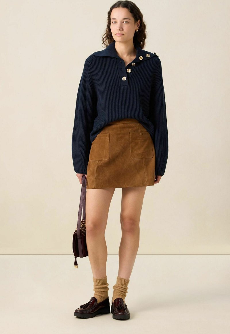 Marineblauwe geribbelde gebreide trui met een brede kraag en gouden knopen, gecombineerd met een bruine suède mini-rok met twee voorzakken. Bruine sokken en schoenen.