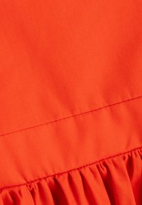 Roter Stoff mit glatter Textur, der am unteren Rand mit einer gerafften Rüsche sowie einer horizontalen Naht versehen ist.