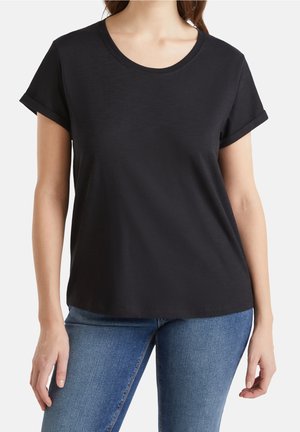 Femme portant un t-shirt noir uni à manches courtes retroussées et un jean bleu, vue du cou aux hanches sur un fond blanc.