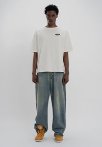 Cream cotton t-shirt with short sleeves and a black oval logo on the chest, paired with light blue baggy jeans and tan work boots.