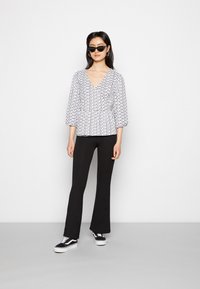 Blusa com padrão em preto e branco, com um decote em V profundo e mangas bufantes, combinada com calças pretas flare e sapatilhas pretas.